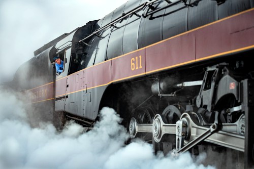 A steam locomotive billows smoke as the engineer leans out of the cab, showcasing vintage train machinery.