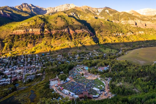 Aerial view of a vibrant outdoor festival surrounded by mountains and greenery, with a crowd gathered in a large open area.