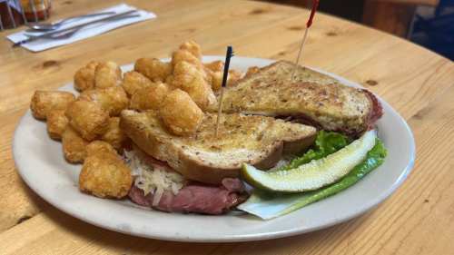 A plate with a sandwich, tater tots, and a pickle, garnished with toothpicks, on a wooden table.