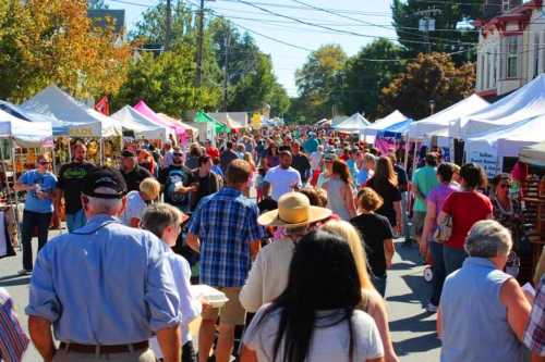 A bustling street fair with crowds of people walking between vendor tents under a clear blue sky.