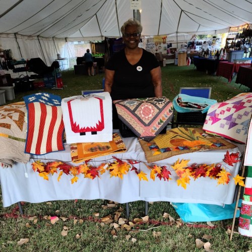 A smiling woman stands behind a table displaying colorful quilts and crafts, with autumn leaves in the foreground.