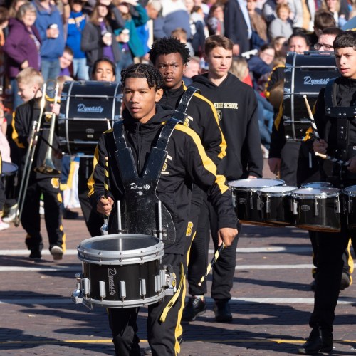 A marching band performs in a parade, featuring drummers in uniforms and a crowd watching in the background.