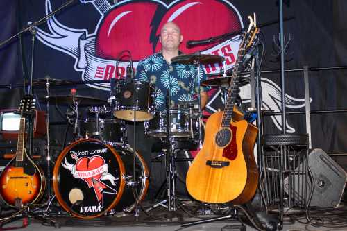 A drummer in a floral shirt sits behind a drum set, with guitars and equipment arranged on stage.