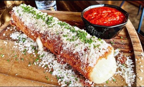 A crispy, cheese-covered corn on the cob topped with herbs, served with a side of red sauce on a wooden platter.