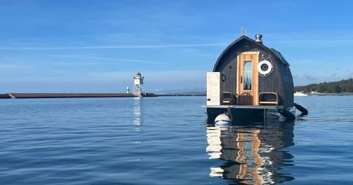 A small, round houseboat floats on calm water, with a lighthouse in the background under a clear blue sky.