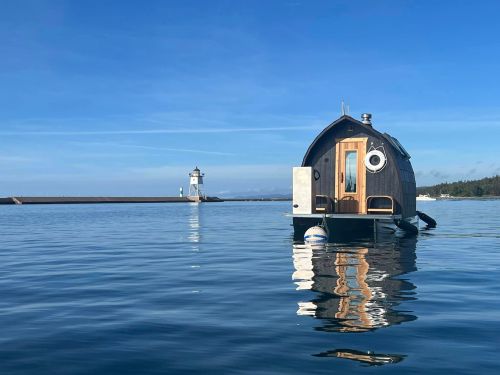 A small, black houseboat floats on calm water, with a lighthouse visible in the distance under a clear blue sky.