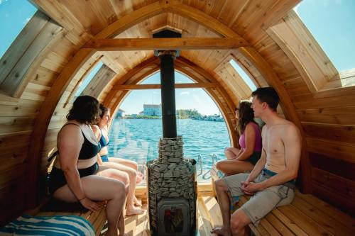 Group of four people sitting inside a wooden sauna, looking out at a lake on a sunny day.
