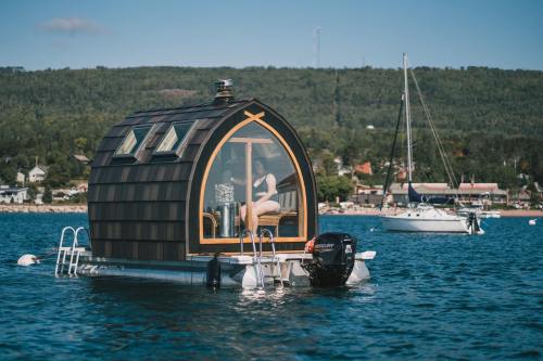 A unique floating cabin on water, with a person inside, surrounded by boats and a scenic landscape in the background.