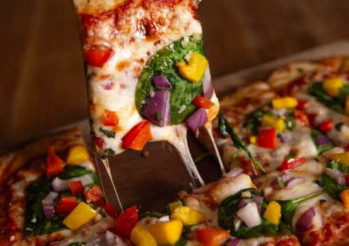 A slice of cheesy pizza topped with spinach, red peppers, onions, and yellow mango, being pulled away from the pie.