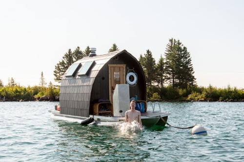 A person swims near a small, modern houseboat surrounded by water and trees under a clear sky.