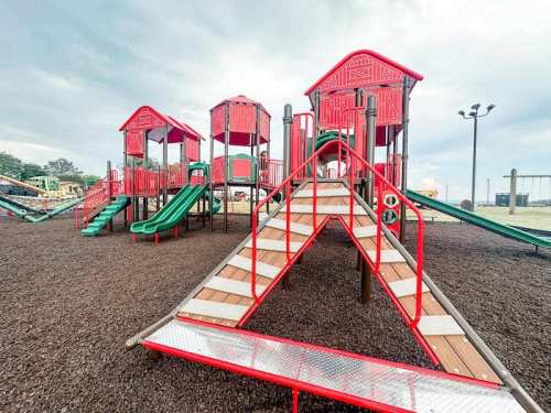 Colorful playground with red structures, slides, and climbing areas, set on a rubber surface under a cloudy sky.