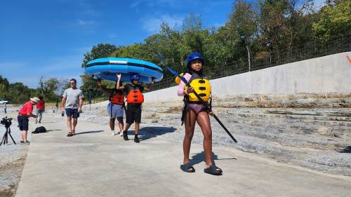 A group of people in life jackets carry inflatable rafts along a path by the water on a sunny day.