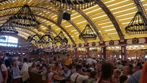 A bustling beer hall filled with people, featuring a band on stage and vibrant yellow-striped ceilings.