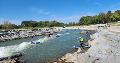 A scenic river with kayakers navigating the water, surrounded by rocky banks and lush greenery under a clear blue sky.