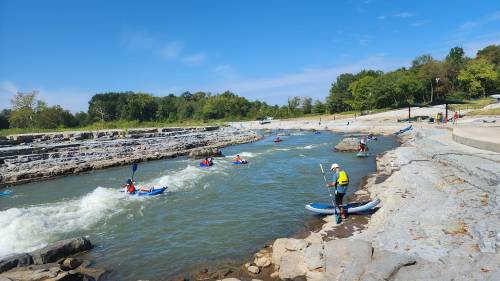 A group of kayakers paddles on a river surrounded by rocky banks and lush greenery under a clear blue sky.