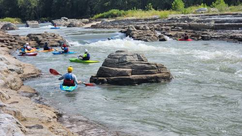 Kayakers navigate a rocky river with rapids, surrounded by lush greenery and large boulders.