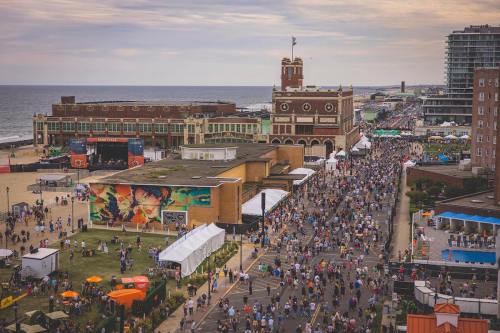 Aerial view of a crowded beach boardwalk event with colorful tents and a historic building by the ocean.
