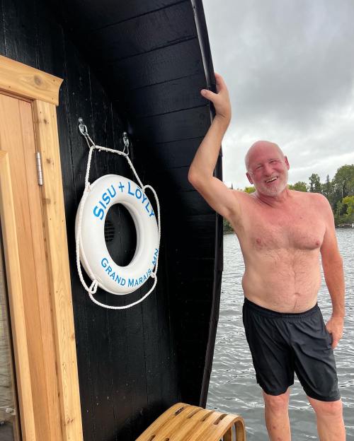 A shirtless man stands by a wooden boat, smiling next to a life ring labeled "SISU LOTTA GRAND MARAIS MN."