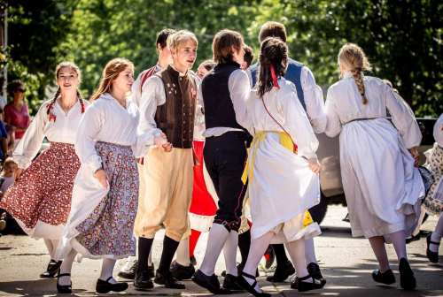 A group of people in traditional costumes dancing together outdoors, surrounded by greenery and sunlight.