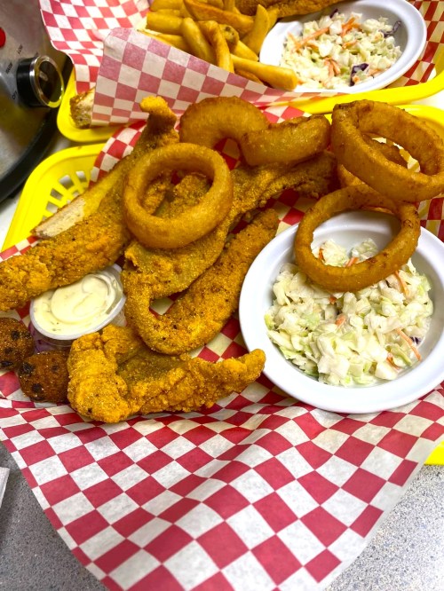 A basket of fried fish fillets, onion rings, coleslaw, and fries served in a red-checkered paper.