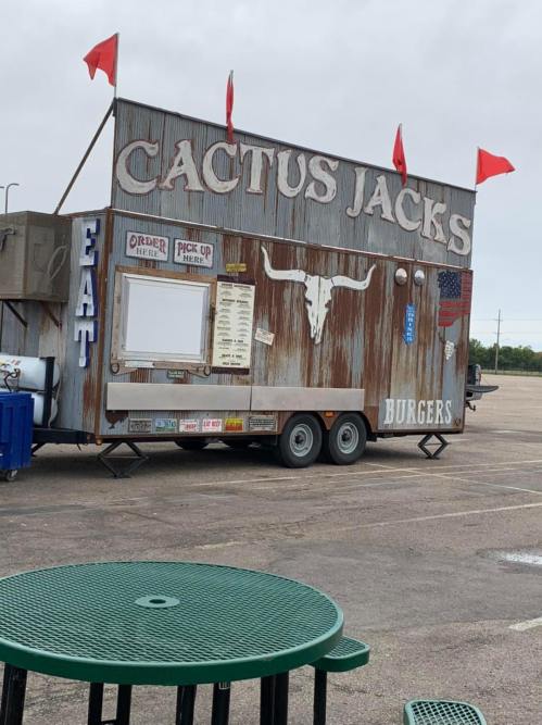 Food truck named "Cactus Jacks" with a rustic design, serving burgers, parked in an empty lot with green tables nearby.