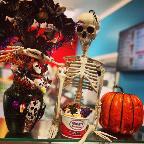 A skeleton sits beside a decorated vase and a pumpkin, with a tub of Hershey's ice cream in a colorful setting.