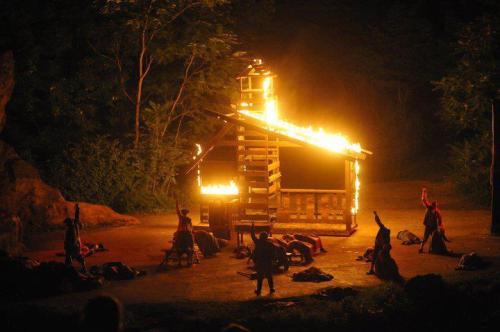 A wooden structure engulfed in flames, with figures nearby, set against a dark forest backdrop.
