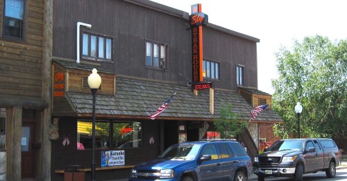 A rustic steakhouse with a wooden exterior, featuring a sign and American flags, surrounded by parked cars.
