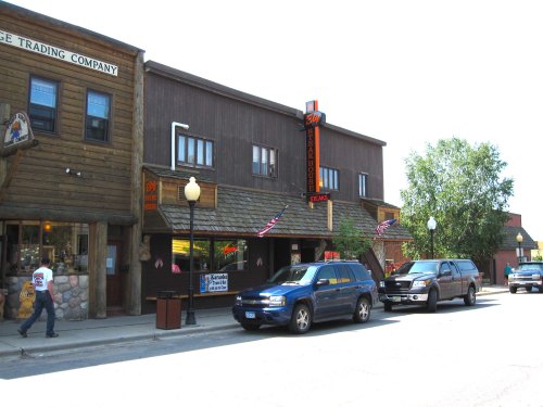 A street view of a rustic town with wooden buildings, parked cars, and American flags displayed.
