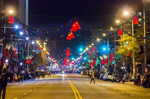 A festive street at night, adorned with colorful lights and decorations, including red bells and garlands.