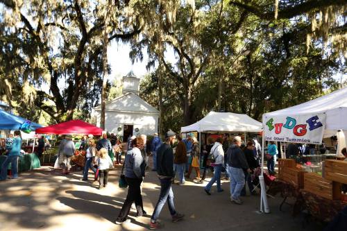 A bustling outdoor market with colorful tents, people walking, and trees draped in Spanish moss.
