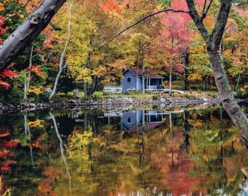 A cozy blue cabin surrounded by vibrant autumn trees, reflecting in a calm lake.