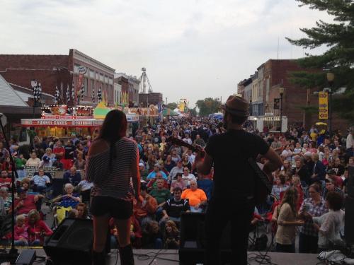 A crowd enjoys a live music performance at a street festival, with food stalls and attractions in the background.