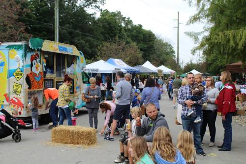 A lively outdoor market scene with food trucks, tents, and families enjoying the day among hay bales and activities.