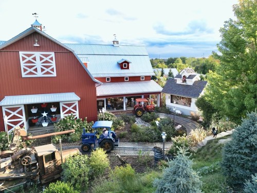 A red barn with a tractor in a garden, surrounded by trees and a quaint building, under a cloudy sky.