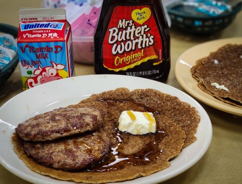 A plate with two sausage patties, syrup, and a dollop of butter on a large pancake, with milk and dessert in the background.
