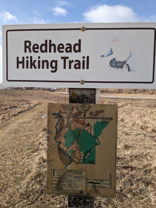 Sign for Redhead Hiking Trail with a map of the trail below, set in a grassy area under a cloudy sky.