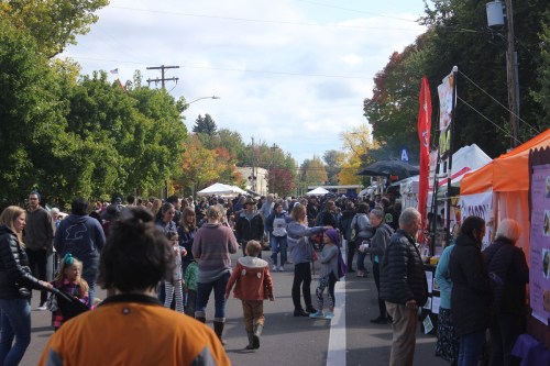 A busy street fair with crowds of people, food stalls, and colorful tents under a partly cloudy sky.