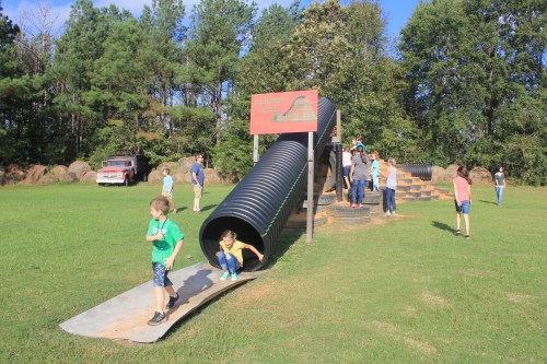 Children playing on a large playground slide and tunnel, with families enjoying the outdoor space in a grassy area.
