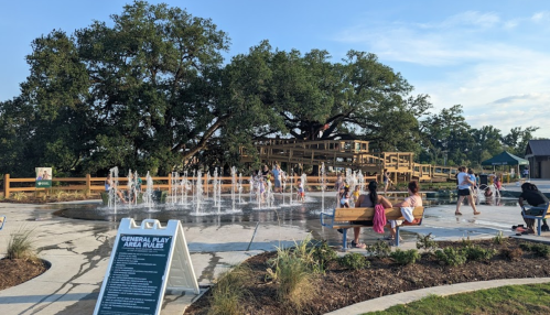 A splash pad with water jets, surrounded by families enjoying a park with a large tree and playground in the background.