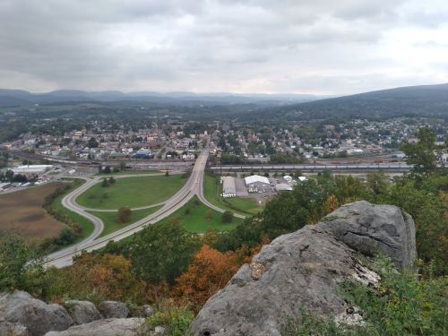 A panoramic view of a small town surrounded by hills, with roads and greenery visible under a cloudy sky.