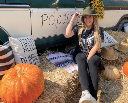 A woman in a hat sits on hay next to pumpkins and a vintage truck, surrounded by decorative pillows and straw hats.