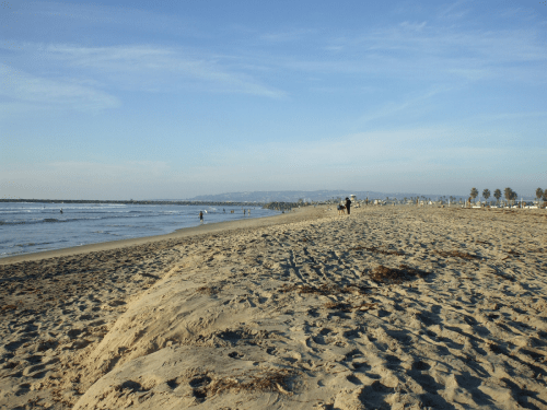 A sandy beach with gentle waves, people walking along the shore, and a clear blue sky in the background.