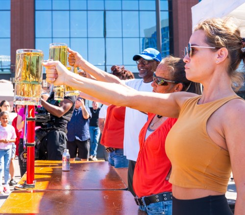 A group of people holding up large beer mugs at an outdoor event, with a crowd in the background.