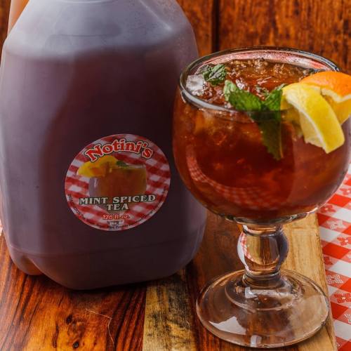 A glass of mint spiced tea with lemon and mint, next to a jug of the same tea on a wooden surface.