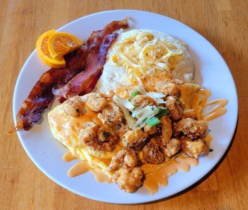 A plate of food featuring an omelet topped with fried shrimp, grits, bacon, cheese, and an orange slice.