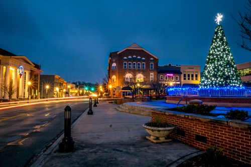 A festive street scene at dusk featuring a decorated Christmas tree and illuminated buildings.