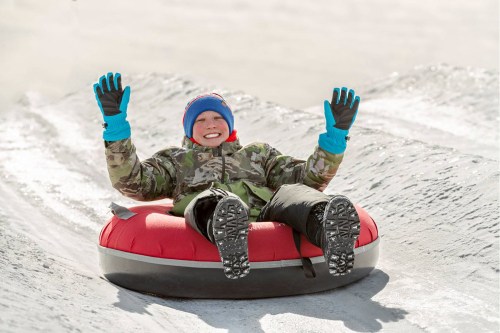 A smiling child in winter gear joyfully rides a red snow tube down a snowy slope, hands raised in excitement.