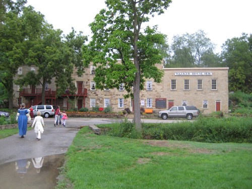 A stone building with multiple windows and a sign, surrounded by trees, with people walking nearby on a path.