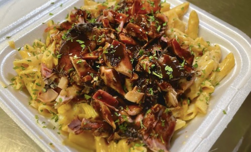 A takeout container filled with pasta topped with shredded meat and drizzled with sauce, garnished with parsley.